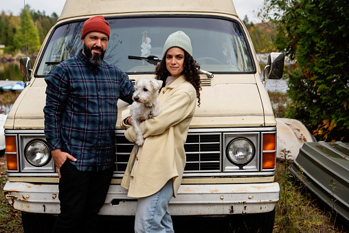 Millennial couple portrait in front of in vintage camper van. They are casually dressed with warm clothes. Dog is a Westie. Lake in the background. Horizontal waist up outdoors shot with copy space. This is part of a series and was taken in Quebec, Canada.