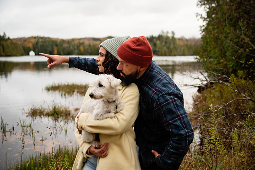 Millennial couple and a dog portrait in front of a lake. They are casually dressed with warm clothes. Dog is a Westie. Lake in the background. Horizontal waist up outdoors shot with copy space. This is part of a series and was taken in Quebec, Canada.