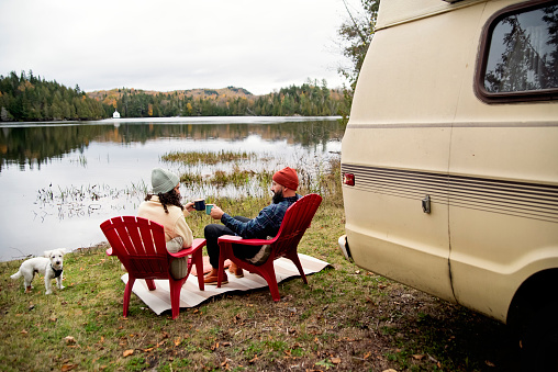Millennial couple enjoying a week-end in vintage camper van. They are looking at the lake view with a drink. They are casually dressed with warm clothes. Dog is a Westie. Horizontal full length outdoors shot with copy space. This is part of a series and was taken in Quebec, Canada.