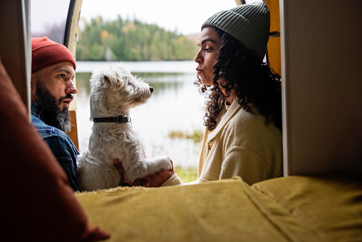 Millennial couple and a dog enjoying a week-end in vintage camper van. They are sitting on the van edge, doors are opened. They are casually dressed with warm clothes. Dog is a Westie. Lake in the background. Horizontal waist up indoors shot with copy space. This is part of a series and was taken in Quebec, Canada.