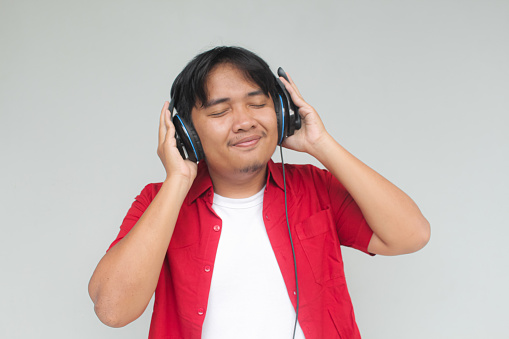 Portrait of attractive Asian man in red shirt enjoying and listening to music using headphone and making dance moves with hands up. Isolated image on gray background.