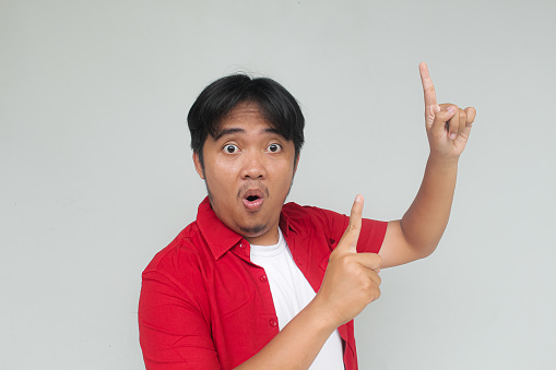 Young asian man in red shirt with happy and wow expression. While pointing up. Isolated on gray background.