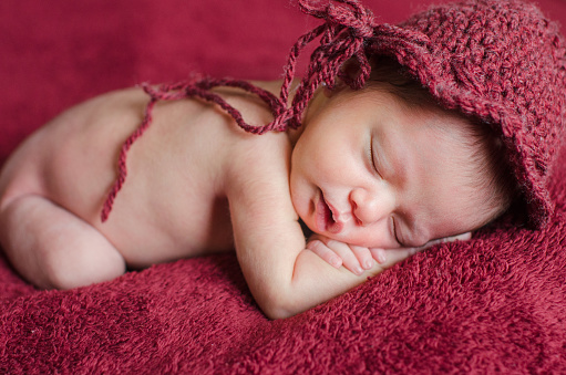 Newborn girl wears a red crochet hat while sleeping in the bed with a red background. Selective Focus