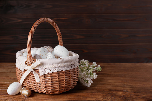 Wicker basket with festively decorated Easter eggs and white lilac flowers on wooden table. Space for text