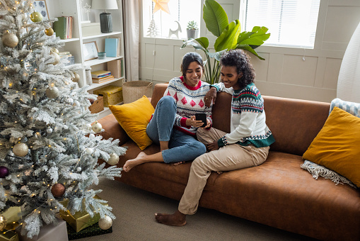 Female gay couple celebrating Christmas at home. They are sitting on sofa and using smart phone for shopping or pleasure.