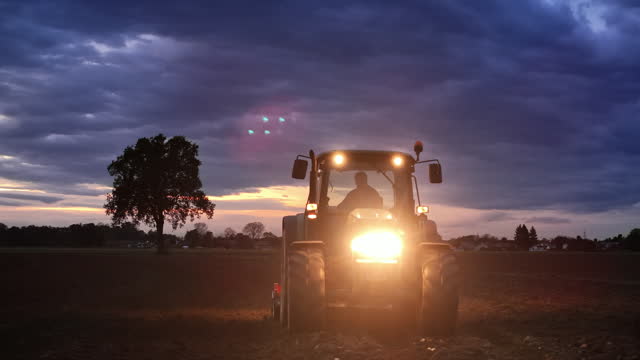 AERIAL Drone Shot of Silhouette Tractor with Lit Headlights Plowing Field under Cloudy Sky in Countryside at Night