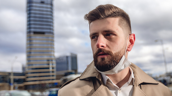 Caucasian guy with face mask after flu. Portrait of bearded handsome man in beige jacket outdoors. Business concept with copy space on the urban street.