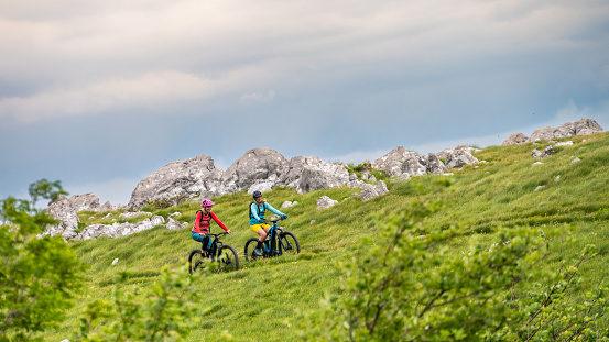 Two women riding electric mountain bikes together at green meadows and cloudscape scenery wide shot side view