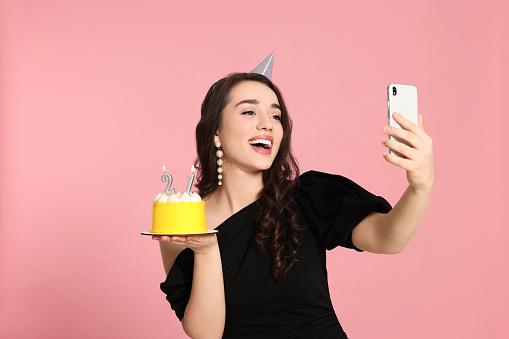 Coming of age party - 21st birthday. Smiling woman holding delicious cake with number shaped candles and taking selfie against pink background