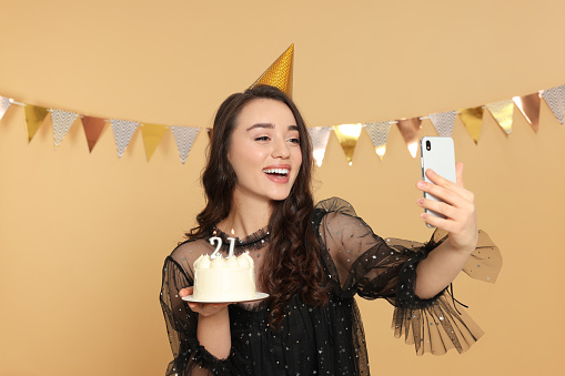 Coming of age party - 21st birthday. Smiling woman holding delicious cake with number shaped candles and taking selfie against beige background