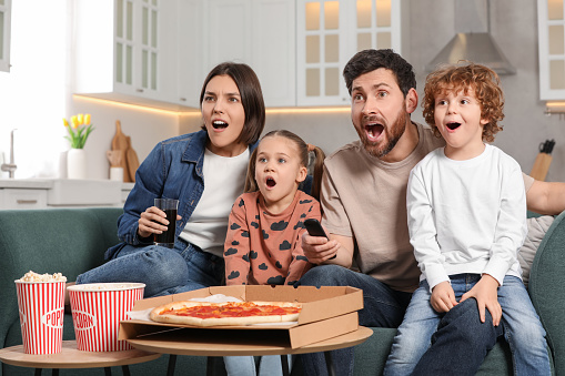 Surprised family watching TV with popcorn and pizza on sofa at home