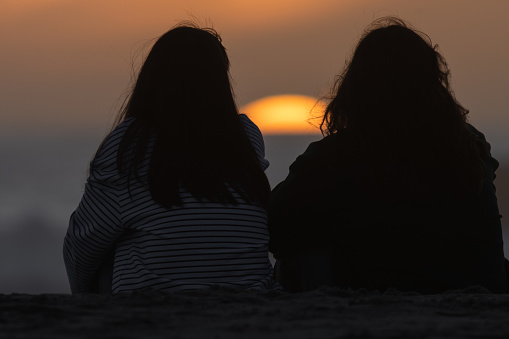 Two girls sitting and watching the sunset. Mid shot