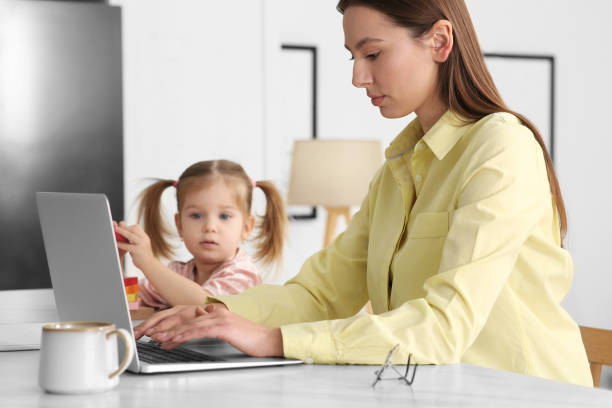 istock Woman working remotely at home. Mother using laptop while daughter playing at desk 1769774139