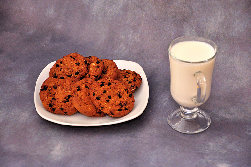 A plate with several oatmeal cookies with chocolate and a glass of hot milk on a gray abstract background. Close-up.