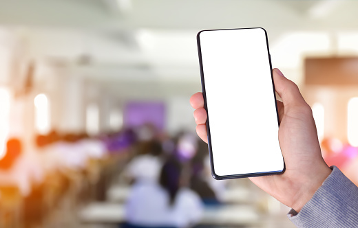 Hand holding blank screen mobile phone with blurred meeting room or classroom, concept for using mobile devices with online learning and conferences.