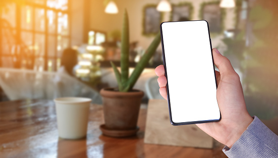 Hand holding white blank screen mobile phone with blurred coffee shop background, concept for online technology with business.