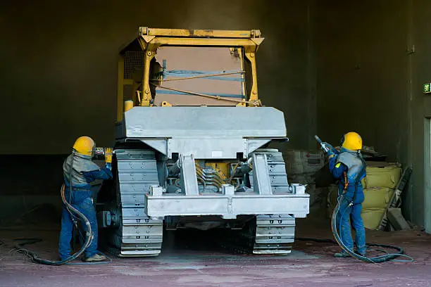 Two men wearing protective clothing and breathing apparatus operate sandblasting equipment to remove paint from a bulldozer in preparation for re-painting.