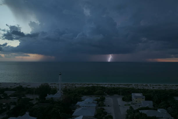 ciudad costera con casas oscuras y faro alto en la orilla del océano con luz parpadeante en la noche de tormenta para la navegación de embarcaciones. tormenta eléctrica con relámpagos sobre el agua de mar que representan un peligro para los barcos - small town america flash fotografías e imágenes de stock