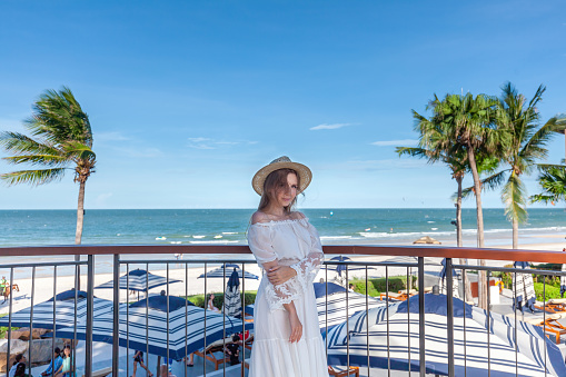 Woman in summer dress observing vast ocean horizon from hotel balcony. Tropical getaway and luxury resort.