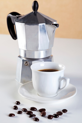 Photo of an Italian Caffettiera or Moka pot with a cup of freshly brewed espresso coffee, a few arabica beans in front. These are also referred to as a Stove pot and are made from aluminium.