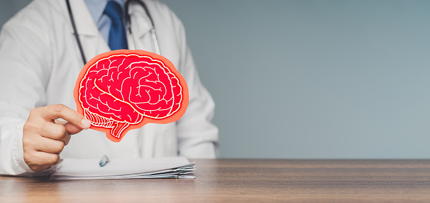 A doctor in a uniform holding a brain symbol made from red paper while sitting at the table in the hospital. Space for text. Medical and healthcare. Brain health and dementia concept