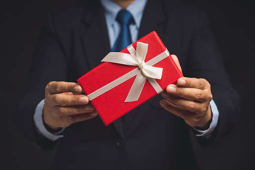 Businessman in a suit holding a red gift box with a white ribbon, giving presents on holiday, bonuses and surprises concept