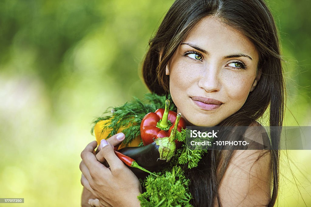 Brunette woman holding vegetables in arms Portrait of young beautiful woman with bare shoulders holding a vegetable - parsley, pepper, eggplant, on green background summer nature. Adult Stock Photo