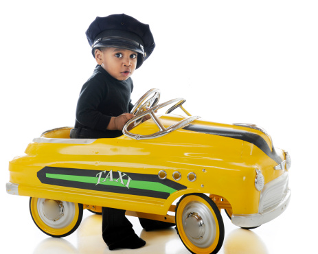 An adorable toddler driving a yellow pedal car while wearing a cab drive's hat.  On a white background.
