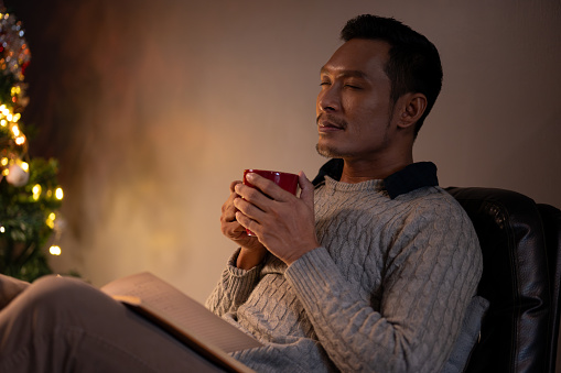 Side view image of a relaxed and calm Asian man in a cosy sweater, eyes closed, smelling his coffee, enjoying his coffee or hot cocoa in a living room on his Christmas night at home alone.