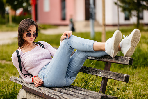 Portrait of a beautiful young woman wearing sunglasses and lying on a park bench in a public park.