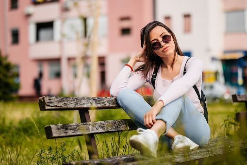 Portrait of a beautiful young woman wearing sunglasses and sitting on a park bench in a public park.