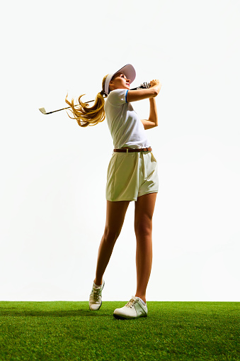 Beautiful, sportive, young blonde girl in white shorts and t-shirt playing golf on grass, teeing off isolated over white background. Concept of sport, hobby, beauty and fashion, relaxation, game