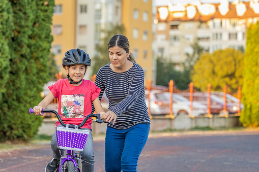 Shot of cheerful young mother who helping her daughter to ride hers bike outside during the day.