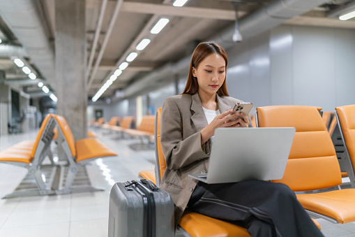 Asian businesswoman working on laptop and checking her smartphone for messages and digital banking transaction while waiting for her flight at the airport, a suitcase by her side. Young woman on business trip. Lifestyle and technology. Business on the go. Digital banking.