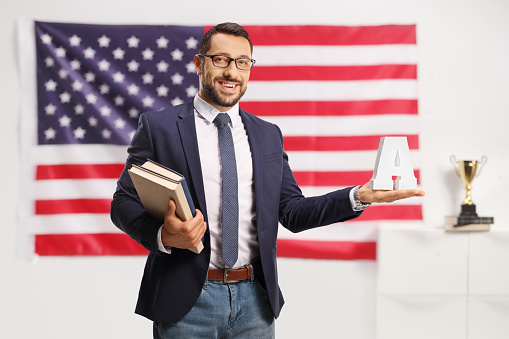 Young man with glasses holding books and the letter A in front of a USA flag