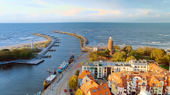 Drone photo captures Koobrzeg's maritime charm, featuring the iconic lighthouse, cerulean sea, turbulent waves, a distant pier, and autumnal hues on the trees.