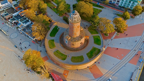 Drone photo captures Koobrzeg's maritime charm, featuring the iconic lighthouse, cerulean sea, turbulent waves, a distant pier, and autumnal hues on the trees.