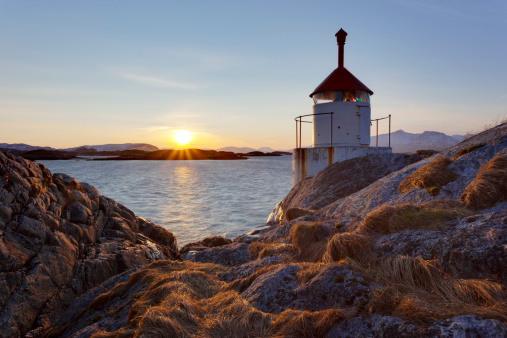 A photograph of a small lighthouse surrounded by orange grasses and rocks near the small fishing village in Northern Norway. The sun is at full height just above the distant mountains and a blue sky fills the upper portion of the frame.