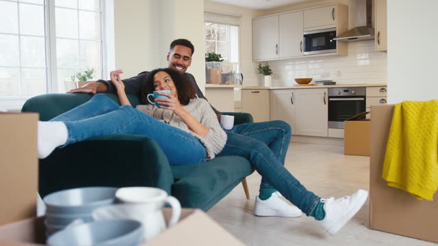 Couple Taking A Break From Unpacking Boxes Relaxing On Sofa With Hot Drink In New Home On Moving Day