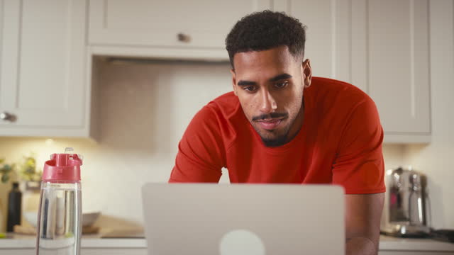 Man Drinking From Bottle Of Water In Kitchen At Home Wearing Fitness Clothing Working On Laptop