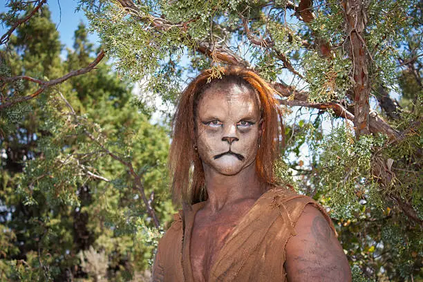 A Wild Lion man stares cautiously at the camera with a background of forest.  This is the fantastic make-up done by Rayce Bird.