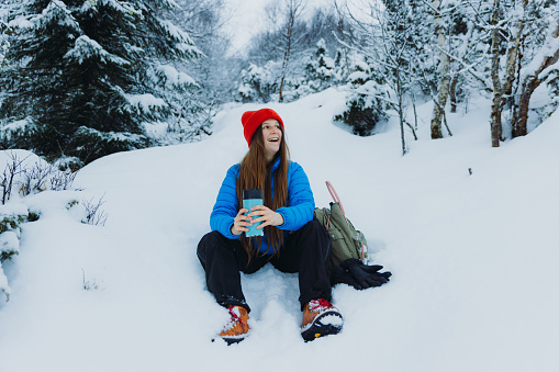 Front View of a smiling female in red hat and blue jacket sitting in the deep snow resting after the hike and enjoying the hot drink in the snowy pine forest in Norway, Scandinavia