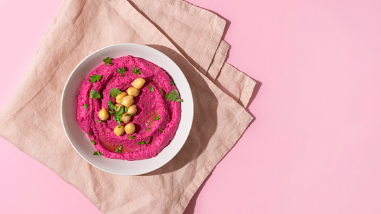 White plate of beetroot hummus decorated with chickpeas on pink towel and background with copy space, top view, hard shadow