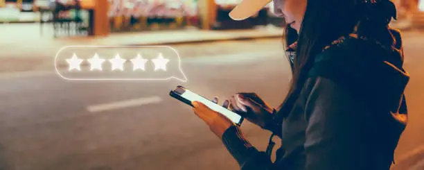 Happy young travel asian woman using mobile phone with graphic of five star icon to give rate of customer service mobile app on street market at night in Bangkok, Thailand