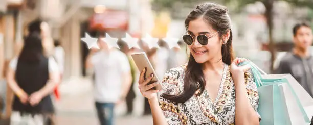 Happy young asian shopping woman using mobile phone with graphic of five star icon to give rate of customer service mobile app on street market in Bangkok, Thailand