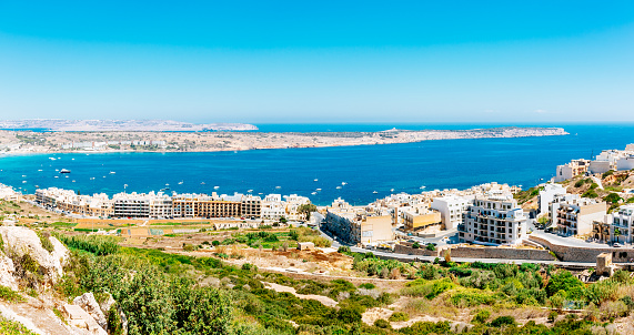 A panoramic photo of a coastal town Mellieha with a view of the sea. The town is built on a cliff and the buildings are white and beige in color. The sea is a deep blue and there are several boats in the water. The sky is a clear blue and there are no clouds. The landscape is green and there are several trees and bushes. The photo is taken from a high vantage.