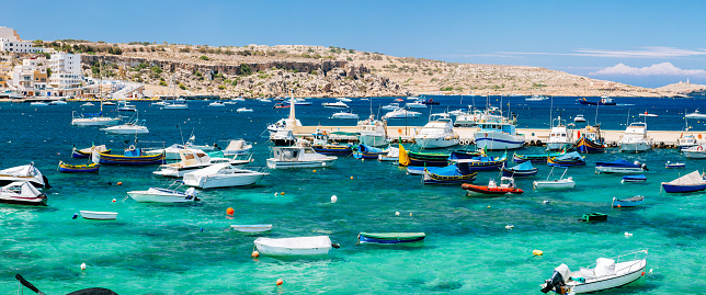A view of the Bugibba bay in Malta, Mediterranean Sea with many boats of different sizes and colors. The boats are anchored near the shore and the water is a deep blue. The sky is clear and the horizon with many houses is visible in the background.