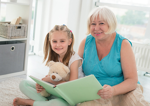 Cute granny reading book to little granddaughter holding toy sitting on floor in playroom