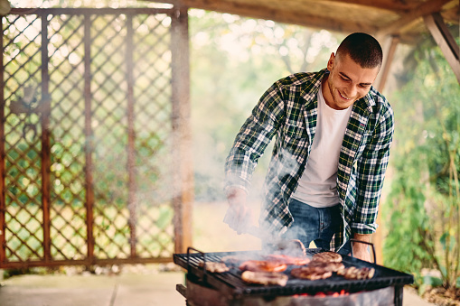 A man making a barbecue outdoors, meat on the grill, smoke all around.