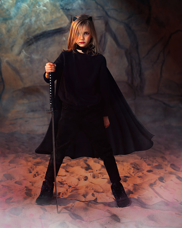 Little girl in ninja superhero costume. Warrior in cosplay with sword for dangerous battle, war or game posing in dark studio cave with smoke. Portrait of girl costume with blade for Halloween